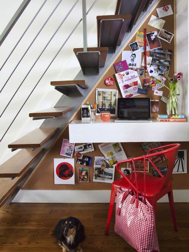 Small workspace under stairs with floating desk and red chair and a small puppy sitting on a hardwood floor. A corkboard is built into the wall and a small laptop is added.